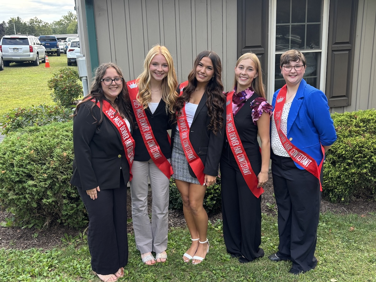 Five young women pose side-by-side outdoors on a grassy lawn in front of a building. They are all smiling at the camera and wearing matching bright red sashes that read "2025 Miss Denver Fair Contestant." They are dressed in professional business-casual attire, including blazers, slacks, and a skirt.