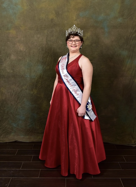 A formal portrait of the 2025 Miss Denver Fair Queen, Annalee McCray. She is a young woman with short brown hair and glasses, smiling warmly while posing against a textured green backdrop. She is wearing a sleeveless, shimmering dark red A-line gown, an intricate silver tiara, and a white sash with a blue border that reads "2025 Miss Denver Fair."