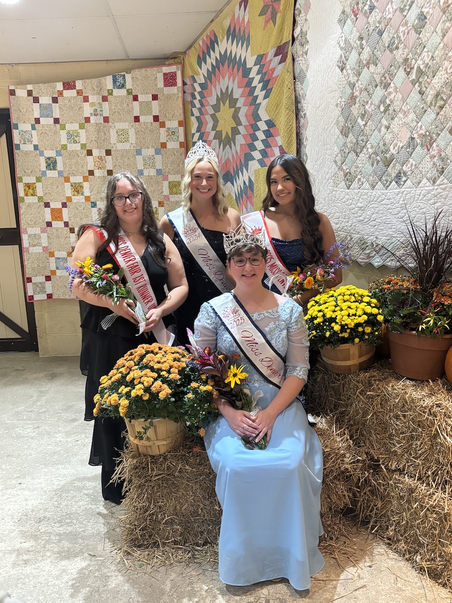 The 2025 Miss Denver Fair Queen and her court pose indoors in front of a rustic display featuring patchwork quilts, hay bales, and potted chrysanthemums. The newly crowned queen is seated in the center wearing a light blue formal gown, a tiara, and a white sash. Standing behind her are three young women in formal gowns holding floral bouquets, including the 1st runner-up and the 2024 Fair Queen.
